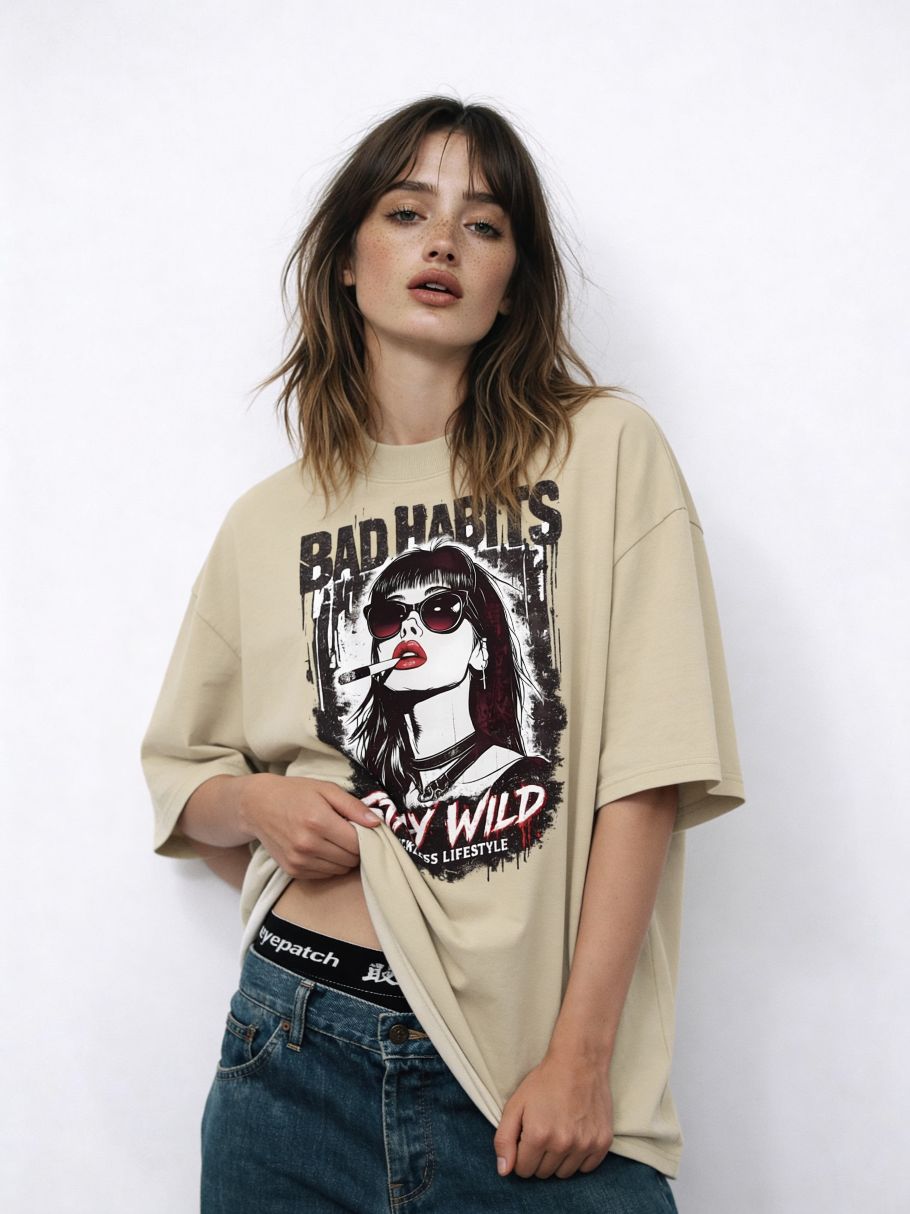 Female fashion model wearing oversized beige “Bad Habits Stay Wild” graphic tee with bold illustration and red lettering, relaxed urban outfit styling