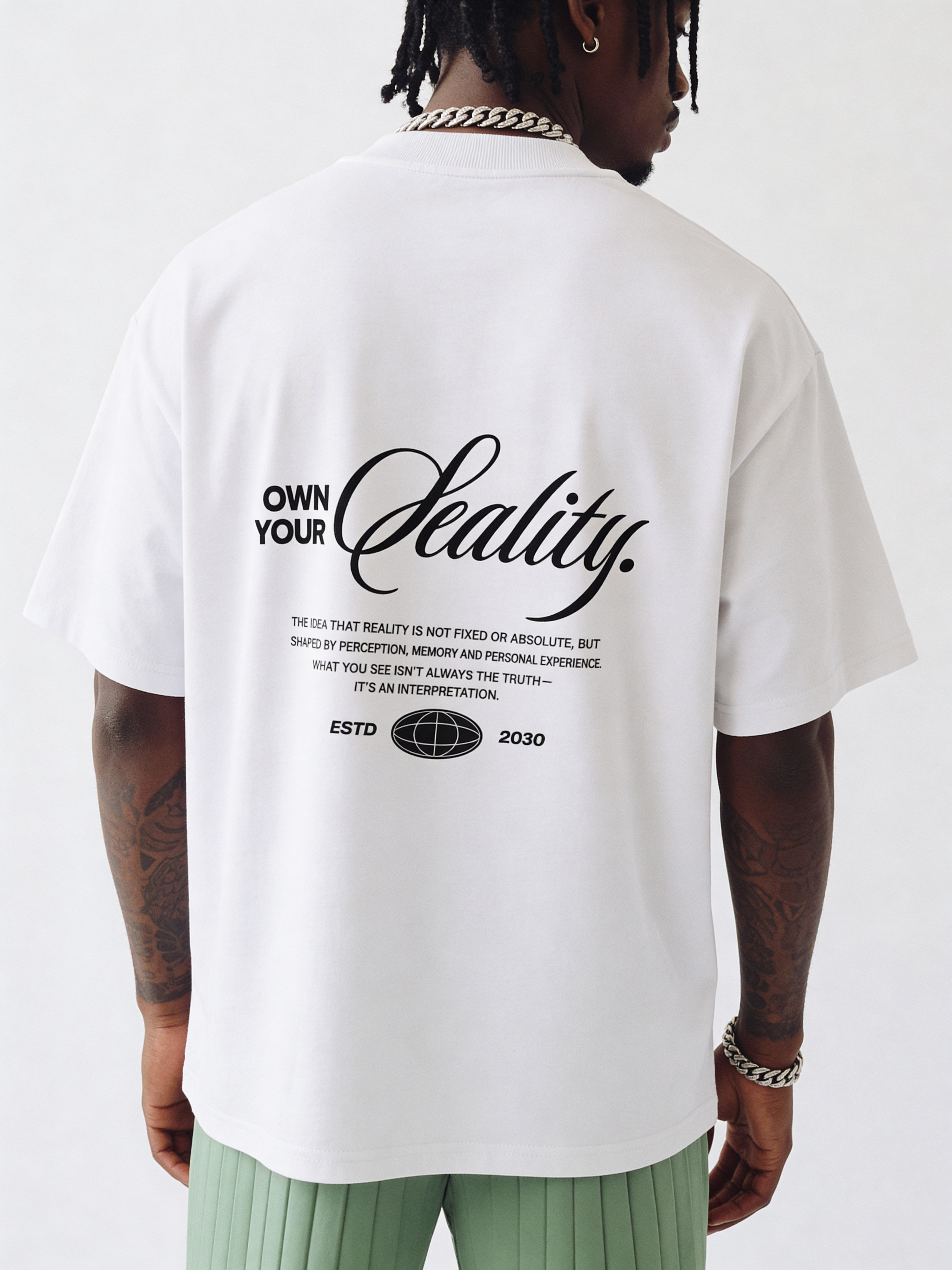 Back view of oversized white streetwear t-shirt with “Own Your Reality” script typography and motivational quote print, worn by male model
