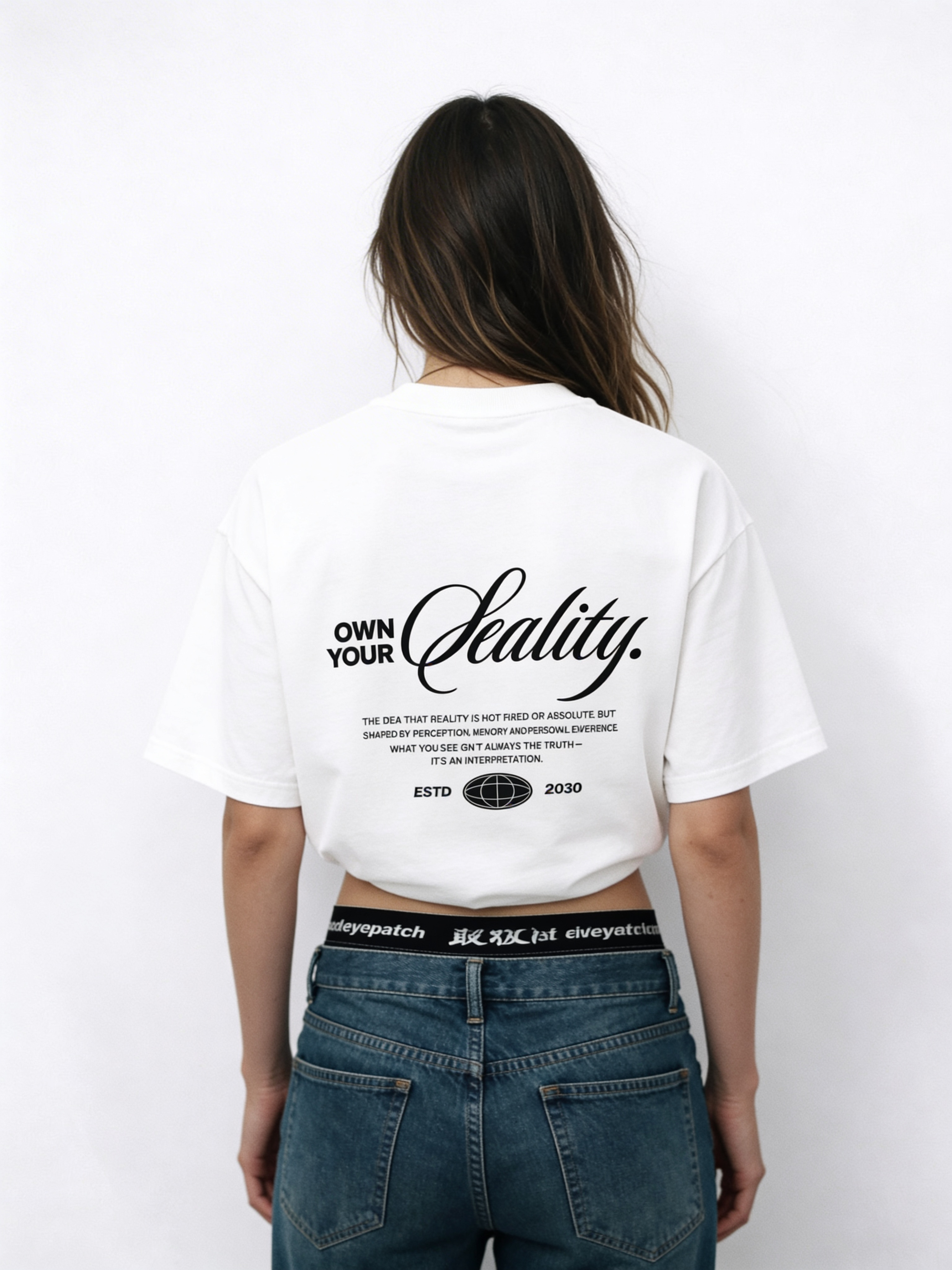 Back view of oversized white “Own Your Reality” streetwear t-shirt with bold script lettering and motivational text graphic, worn by female model with jeans