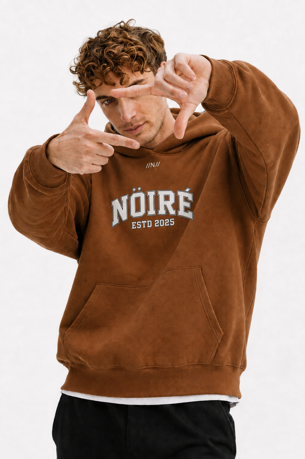 Male model wearing brown oversized Nöiré ESTB 2025 hoodie with kangaroo pocket, close-up lifestyle view, streetwear fashion