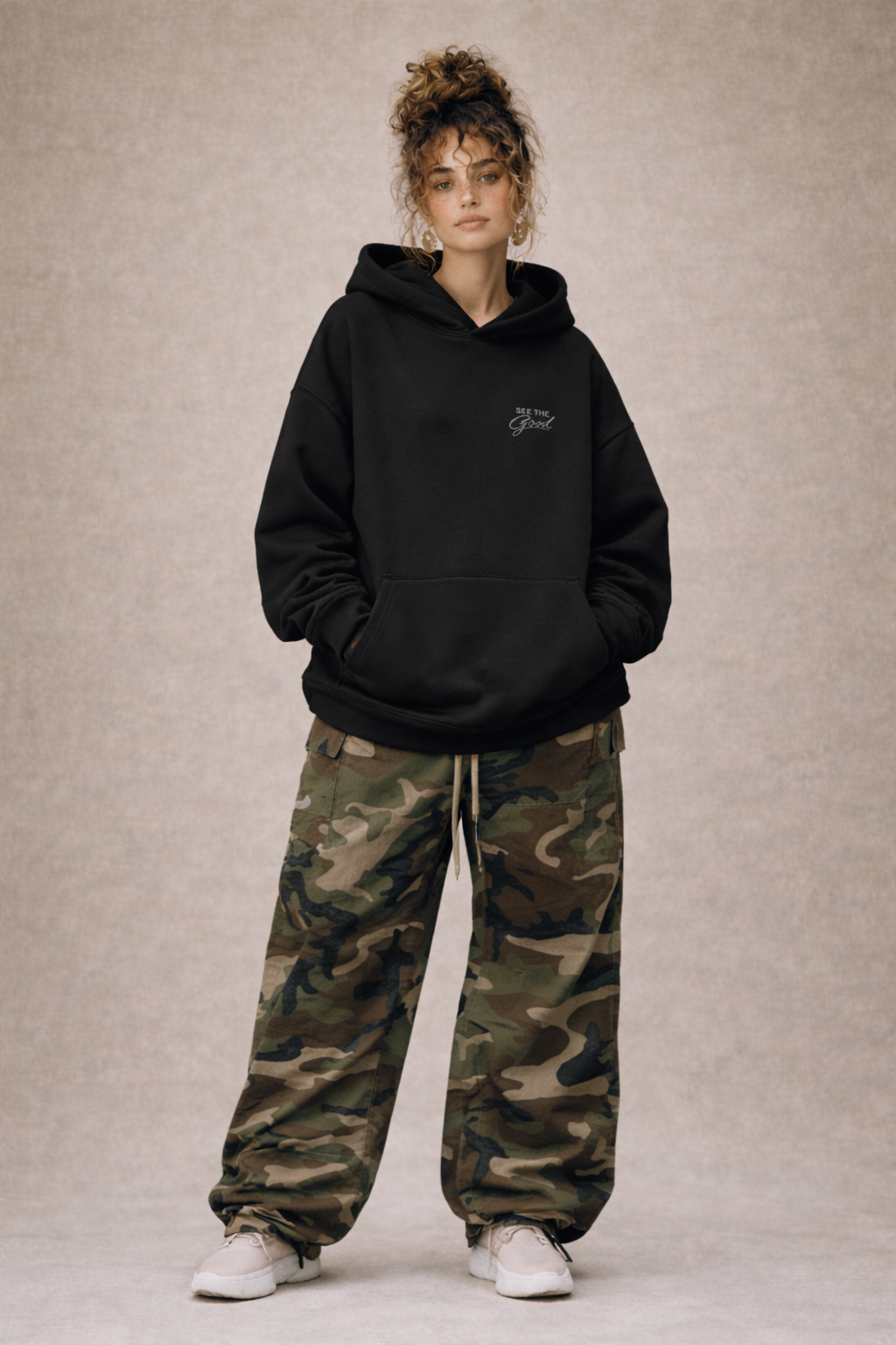 Full body view of woman wearing black oversized hoodie with subtle “See The Good” chest print, styled with camouflage cargo pants, modern women’s streetwear fashion.