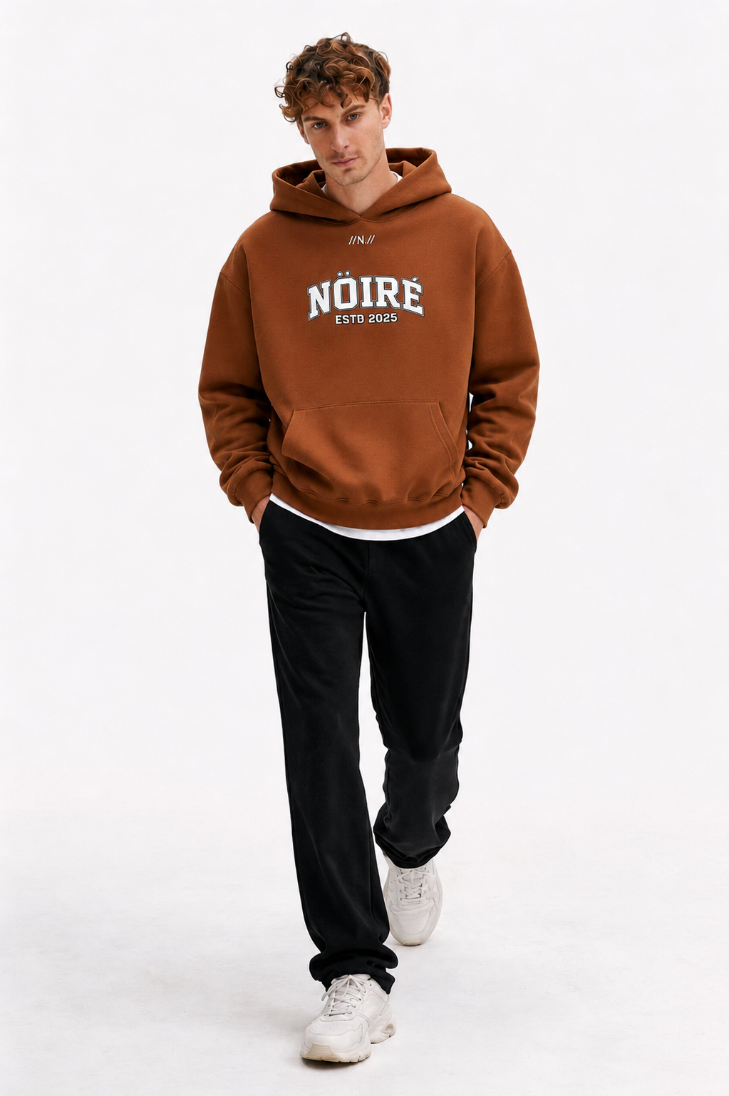 Male model walking in brown oversized Nöiré ESTB 2025 logo hoodie, full-body streetwear outfit