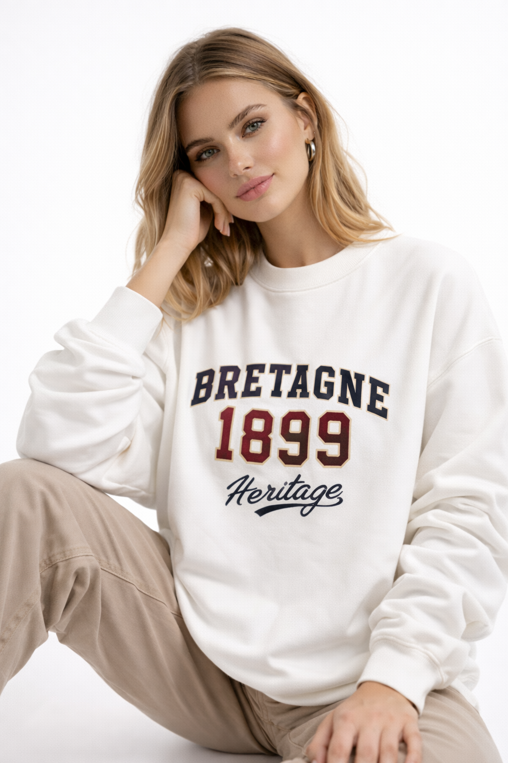Close-up of woman wearing cream oversized sweatshirt with bold “Bretagne 1899 Heritage” retro varsity print, minimalist neutral streetwear style.