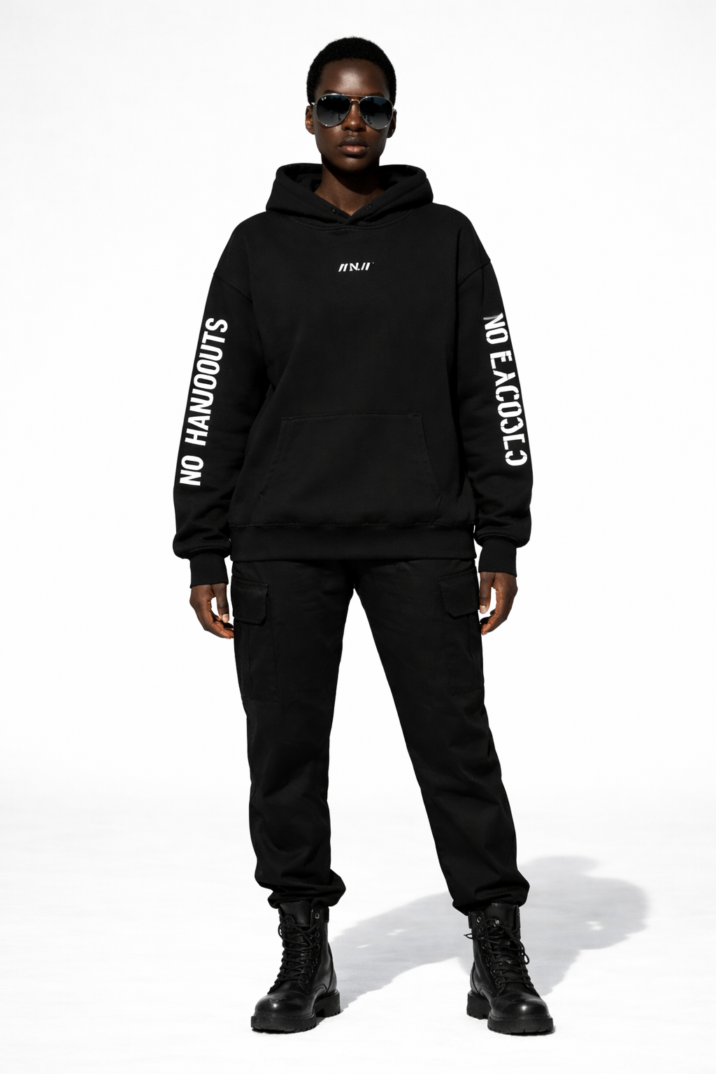Female model front full-body wearing black oversized hoodie with “//N.//” chest logo and bold sleeve prints, black cargo pants and boots, minimal studio background.