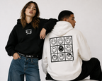 female model leaning pose wearing black cropped graphic hoodie while male model seated back view wearing oversized cream hoodie with smiley artwork streetwear fashion
