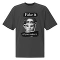 Faded Black unisex t-shirt front view with illustrated face graphic and “Fake it til you make it” slogan, modern streetwear tee